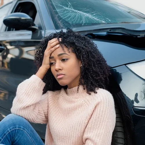 Young woman leans against her damaged car, holding her head in pain after an accident